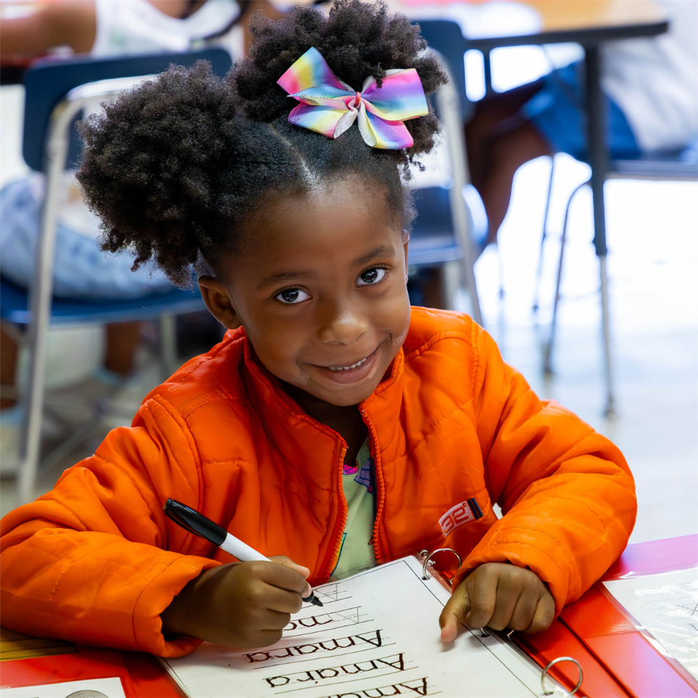 a young girl in an orange jacket looks a the camera while writing